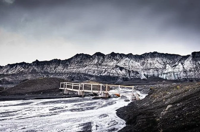 A footbridge over a glacial river below an ash-covered glacier